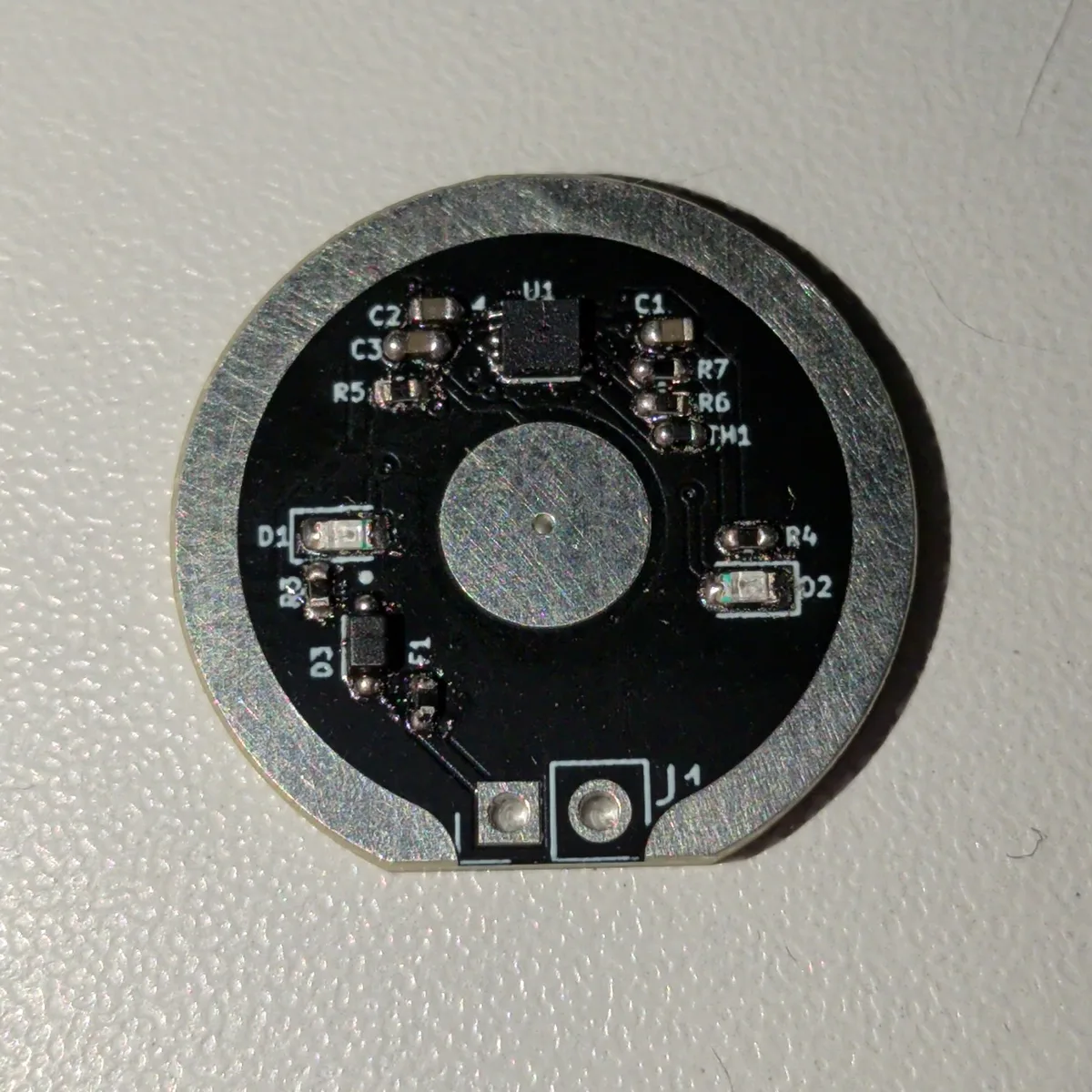 The assembled V1.0 prototype board, front side: a black ring PCB with a charger chip, two small LEDs, a fuse, a diode and a thermistor around a round contact pad.